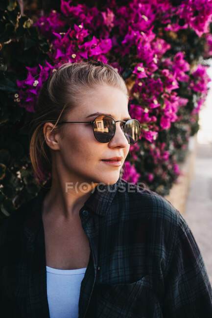 Bella signorina in abito alla moda e occhiali da sole guardando lontano mentre in piedi vicino cespuglio con bellissimi fiori sulla strada — Foto stock