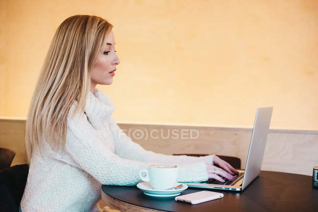 Signora che utilizza il computer portatile a tavola con tazza di bevanda e smartphone — Foto stock