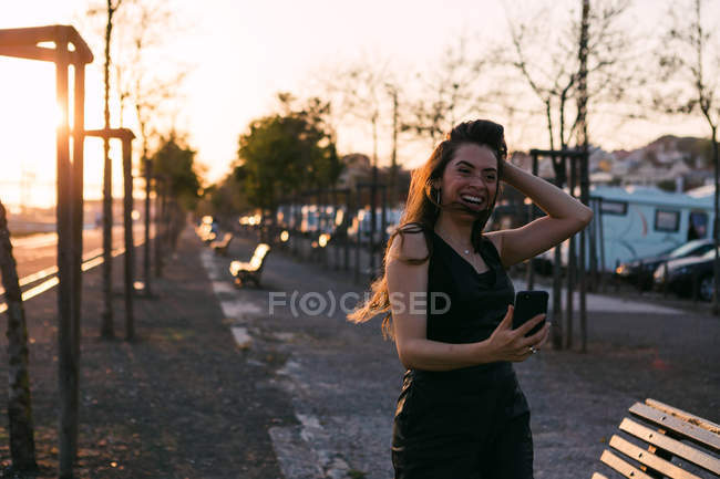 Mujer joven riendo en ropa negra con la mano en el pelo tomando selfie en la calle al atardecer - foto de stock