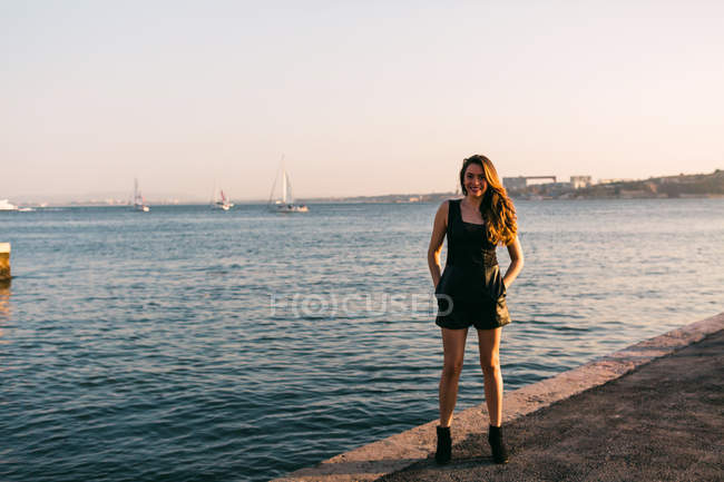 Jovencita sonriente en vestido negro y botas con las manos en los bolsillos de pie en el terraplén cerca de la superficie del agua con barcos al atardecer - foto de stock