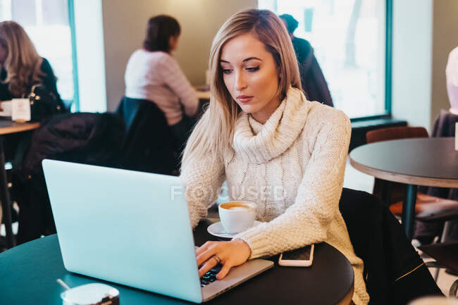 Woman using laptop at table with cup of drink and smartphone — Stock Photo