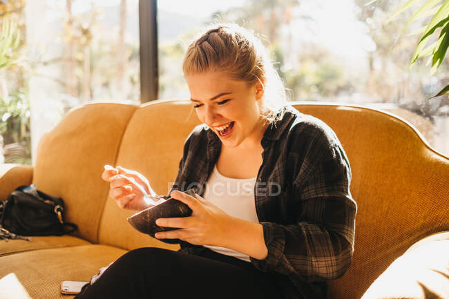 Hübsche junge Frau mit Schüssel und Löffel lächelt, während sie auf einer bequemen Couch am Fenster sitzt — Stockfoto