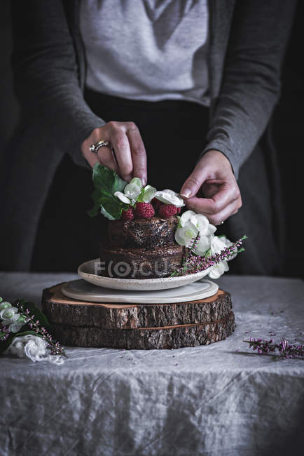 Female hands decorating chocolate cake decorated with raspberries and flowers on plate on wooden stand — Stock Photo