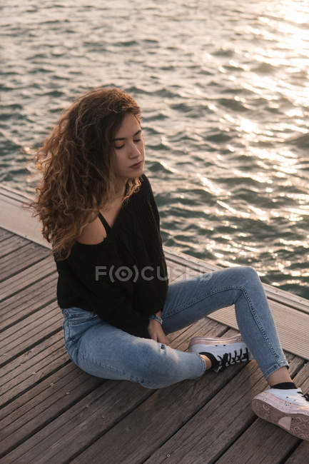 Charming young female with eyes closed sitting on wooden pier near water — Stock Photo