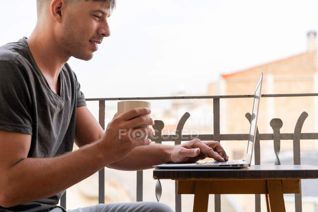 Красивий чоловік з чашкою кави і використовує ноутбук на балконі — стокове фото