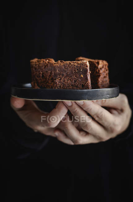 Mains humaines tenant une assiette avec des morceaux de brownie au chocolat sur fond sombre — Photo de stock