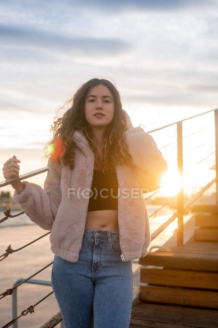 Young lady in stylish outfit touching curly hair and looking at camera while leaning on railing on blurred background of pier and water at sunset — Stock Photo