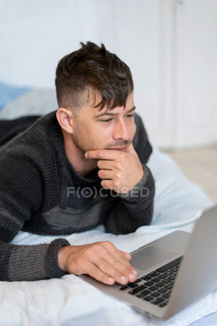 Випадковий молодий чоловік в светрі лежить на ліжку і дивиться, як ноутбук проводить час вдома — стокове фото