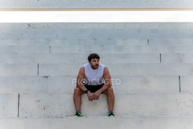 Красивый взрослый парень в спортивной одежде смотрит в сторону, сидя на бетонной лестнице во время тренировки на улице — стоковое фото