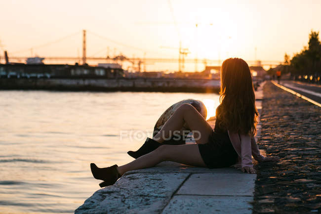 Verträumte sinnliche junge Frau sitzt bei Sonnenuntergang auf einem Damm nahe der Wasseroberfläche — Stockfoto