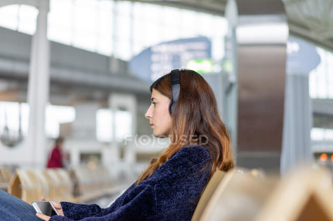Vista lateral atraente senhora segurando celular, ouvindo música e sentada no banco na sala de espera no Porto, Portugal — Fotografia de Stock