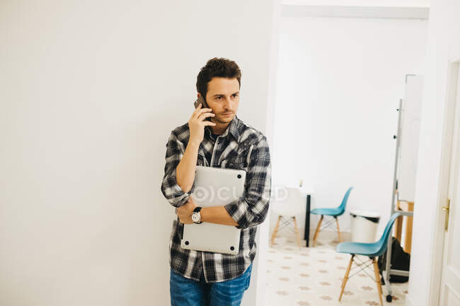 Concentrated guy using mobile phone and holding laptop near white wall and light room with chairs — Stock Photo