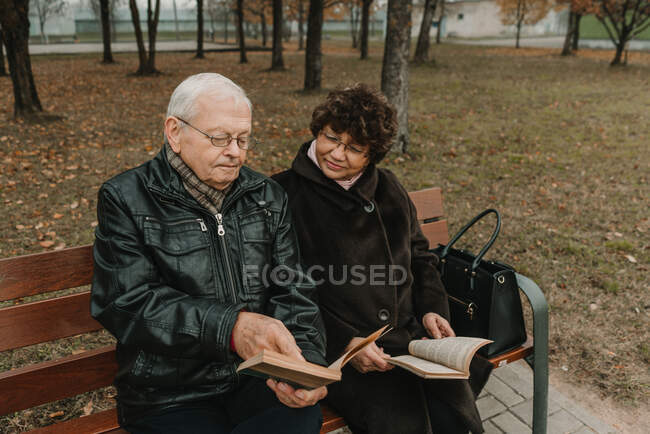 Літній чоловік і жінка сидять на лавці в осінньому парку і читають цікаві книги разом — стокове фото