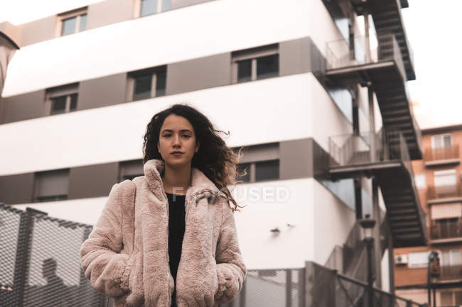 Porträt einer jungen Frau mit lockigem Haar und Händen in Taschen, die in der Nähe eines modernen Gebäudes an der Stadtstraße steht — Stockfoto