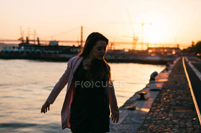 Jovem mulher caminhando no aterro perto da superfície da água ao pôr do sol — Fotografia de Stock