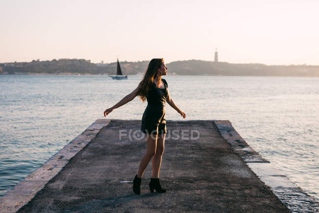 Весела жінка в чорному одязі і чоботи танцюють на набережній біля поверхні води з яхтою на заході сонця — стокове фото