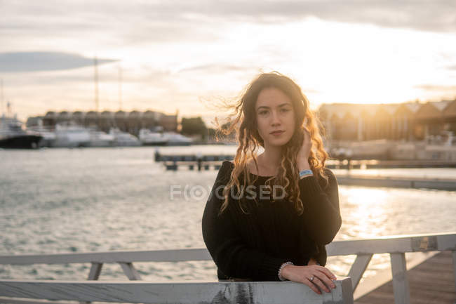 Junge Dame in stylischem Outfit berührt lockiges Haar und blickt in die Kamera, während sie sich auf Geländer auf verschwommenem Hintergrund von Pier und Wasser stützt — Stockfoto