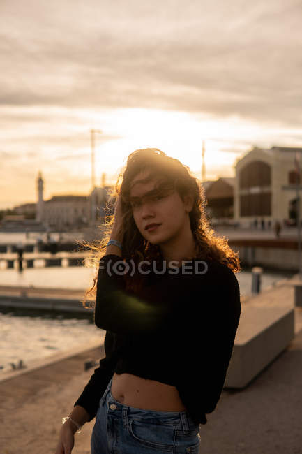 Preciosa joven con el pelo en la cara de pie cerca del agua en el terraplén de la ciudad durante la puesta del sol - foto de stock