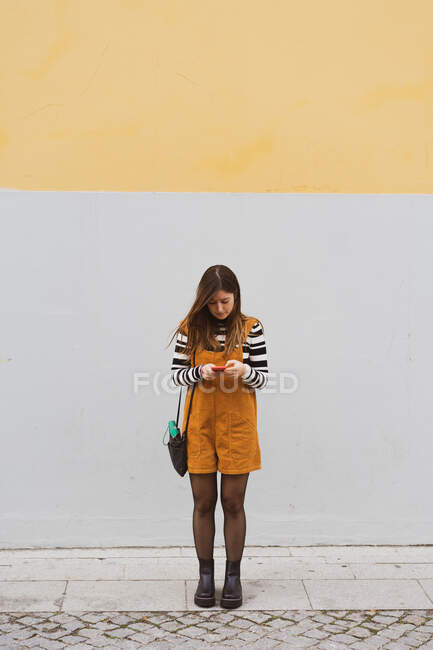 Senhora de camisola e macacão com bolsa a navegar no telemóvel perto da parede cinzenta e castanha na rua do Porto, Portugal — Fotografia de Stock
