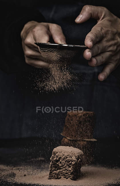 Human hands powdering pieces of chocolate brownie with cocoa on dark background — Stock Photo