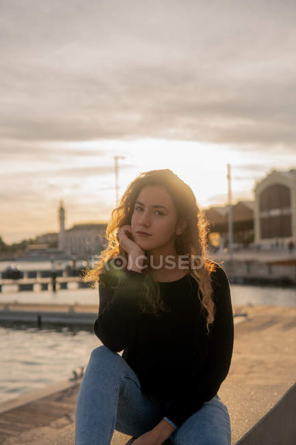 Belle jeune femme soutenant la tête et regardant la caméra tout en étant assis près de l'eau sur le remblai de la ville pendant le coucher du soleil — Photo de stock