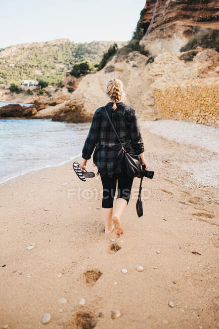 Rückansicht einer jungen Barfußdame mit Fotokamera, die auf Sand in der Nähe von Meerwasser spaziert — Stockfoto