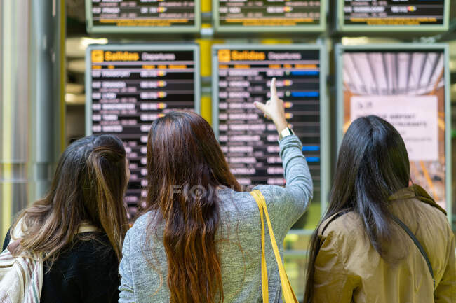 Вид сзади длинноволосых женщин, показывающих пальцем на дисплее информации о рейсах в аэропорту Порту, Португалия — стоковое фото
