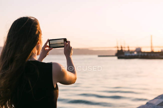 Vista trasera del barco de tiro de mujer en el teléfono inteligente en el terraplén cerca del agua al atardecer - foto de stock