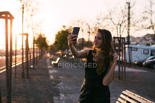 Привабливою леді в чорне плаття з боку у волоссі, беручи selfie на вулиці на заході сонця — стокове фото
