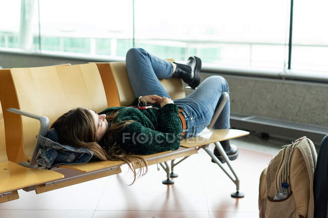 Señora vista lateral descansando en el banco en la sala de espera del aeropuerto cerca de la ventana en Oporto, Portugal - foto de stock