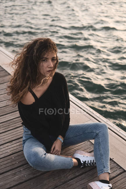 Junge Frau blickt in die Kamera, während sie auf einem Holzsteg am Wasser sitzt — Stockfoto