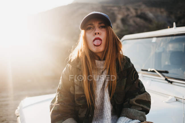 Attractive red haired woman showing tongue, looking at camera and sitting on off-roader near hills — Stock Photo
