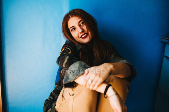 Sorridente donna attraente seduta vicino al muro blu — Foto stock