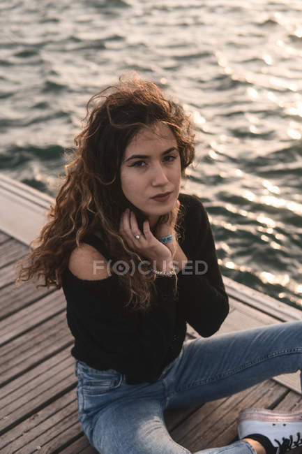 Молода жінка дивиться на камеру, сидячи на дерев'яному пірсі біля води — стокове фото