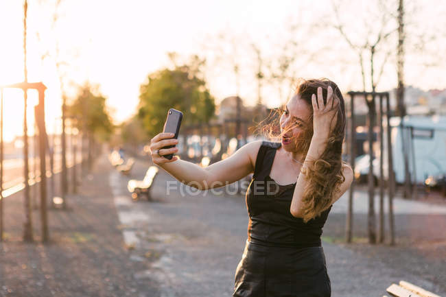 Приваблива дама в чорній сукні з рукою в волоссі бере селфі на вулиці на заході сонця — стокове фото