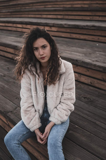 Attraktive junge Frau in stylischem Outfit blickt in die Kamera, während sie auf einer Holztreppe in der Stadtstraße sitzt — Stockfoto