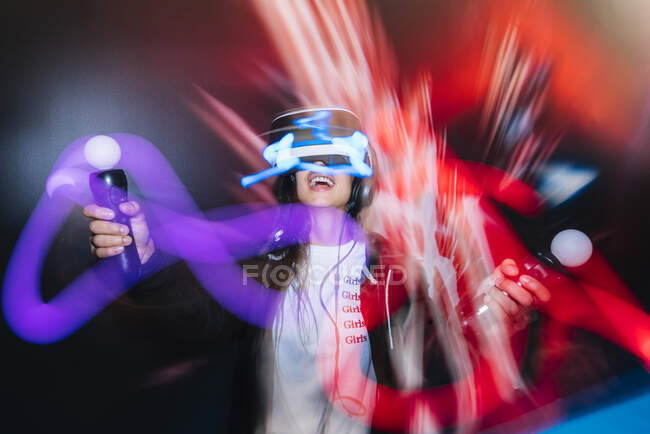 Borroso tiro de larga exposición de la mujer feliz en gafas VR jugando videojuego con controladores en la oscuridad - foto de stock