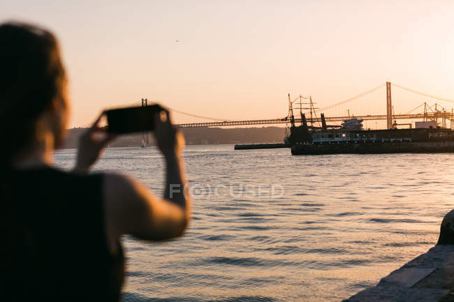 Вид ззаду жінка стріляє човном на смартфон на набережній біля води на заході сонця — стокове фото