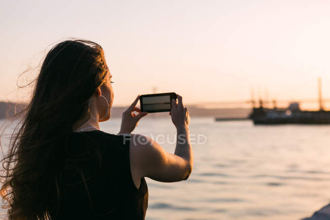 Вид ззаду жінка стріляє човном на смартфон на набережній біля води на заході сонця — стокове фото