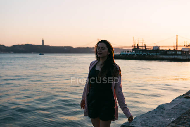 Мрійлива молода дівчина, що йде на набережній біля поверхні води на заході сонця — стокове фото