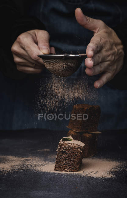 Human hands powdering pieces of chocolate brownie with cocoa on dark background — Stock Photo
