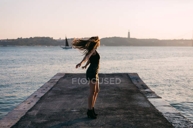 Cheerful woman in black wear and boots dancing on embankment near water surface with yacht at sunset — Stock Photo