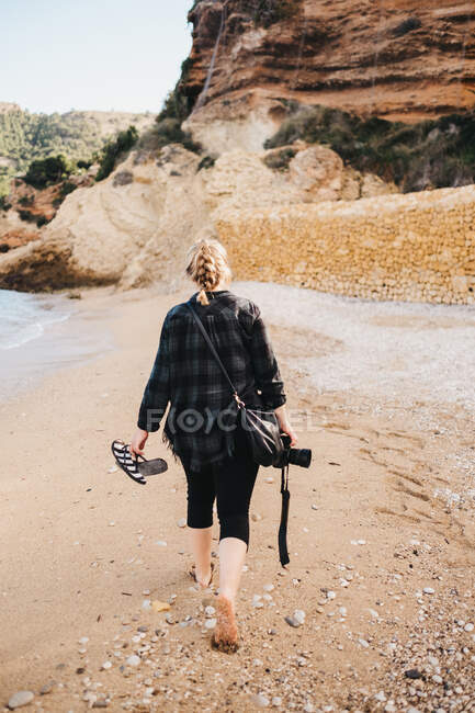 Back view of young barefoot lady with photo camera walking on sand near sea water — Stock Photo