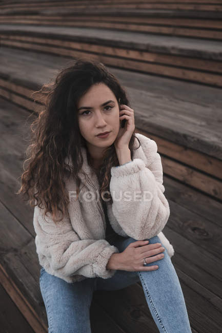 Attraente giovane donna in abito elegante guardando la fotocamera mentre seduto su scale di legno sulla strada della città — Foto stock