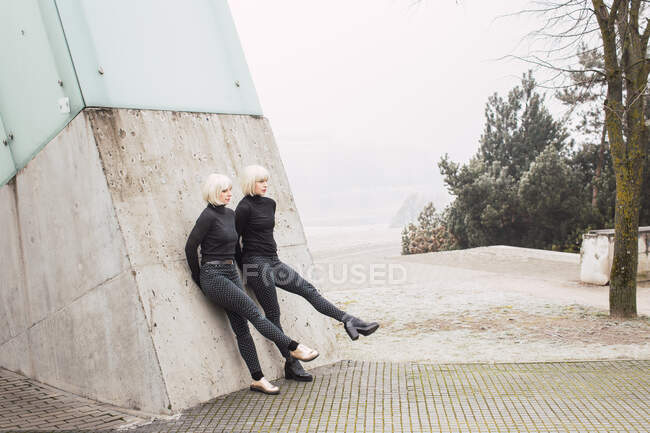 Attractive young women in dark wear stretching out legs and leaning on wall on street near trees in Lithuania — Stock Photo