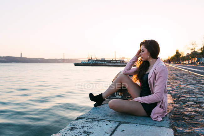 Sonhador sensual jovem senhora sentado em aterro perto da superfície da água ao pôr do sol — Fotografia de Stock