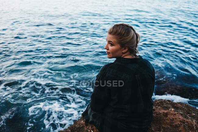 Vista laterale della femmina scalza in abito casual seduta sulla costa rocciosa vicino al mare ad Altea, Spagna — Foto stock