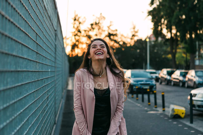 Смеющаяся молодая женщина, идущая по улице возле машин на закате — стоковое фото