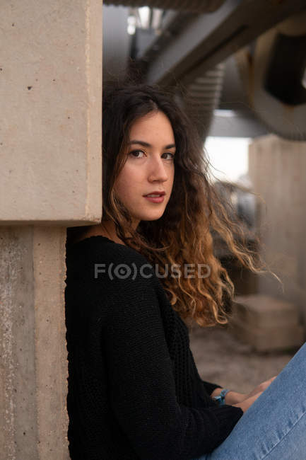 Charmante jeune femme avec les cheveux bouclés regardant la caméra tout en étant assis près du mur de bâtiment en béton — Photo de stock