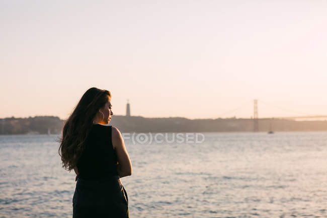 Jovem senhora no desgaste preto em pé no aterro perto da superfície da água ao pôr do sol e sonhando — Fotografia de Stock